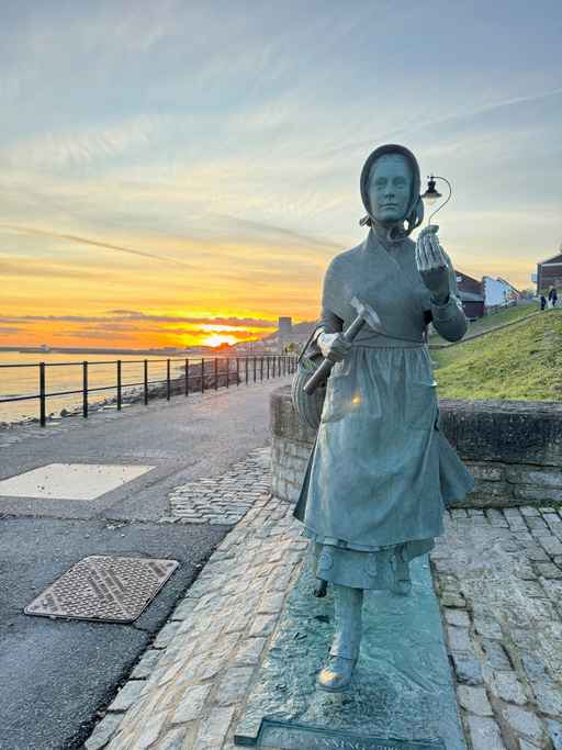 Bronze statue of Mary Anning by the sea at sunset.