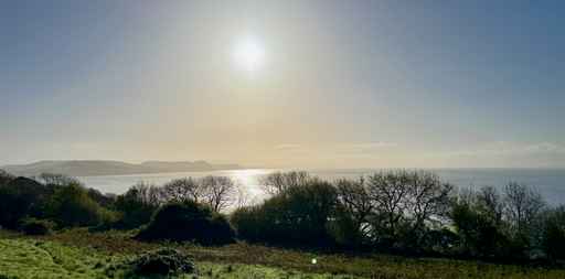 Bright sun over a quiet sea bordered by tree-lined hills.