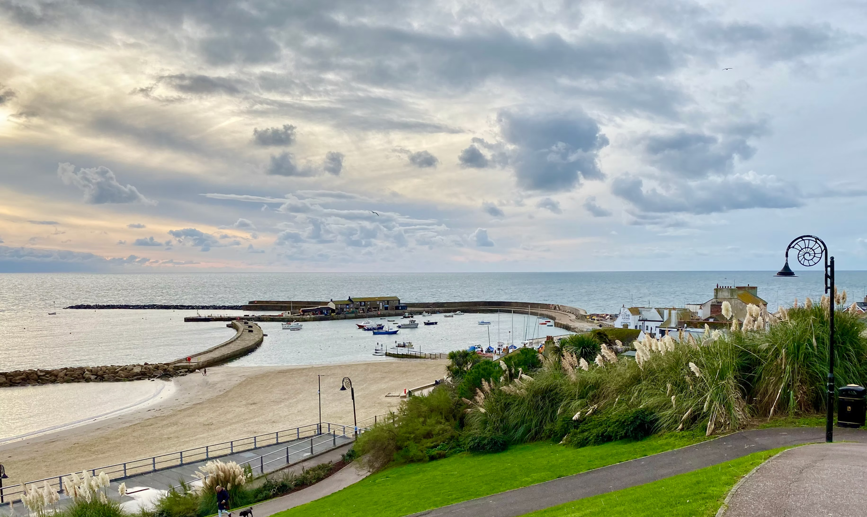 View over the sandy beach and harbour at Lyme Regis, with boats moored behind the Cobb under a cloudy sky during early sunset.